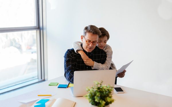 father working on a computer in his home office with his son hugging him from behind
